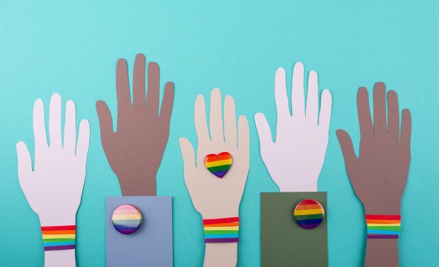 Hands of different complexions all reading up, wearing varied LGBTQ identity pins and wristbands
